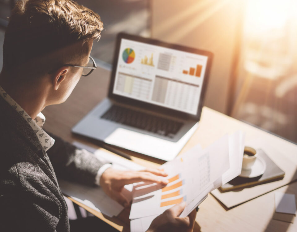 man looking at financial graphs papers spreadsheets on laptop in amber light