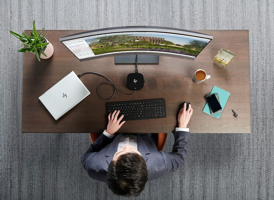 overhead man in suit sitting at wood desk HP laptop curved monitor